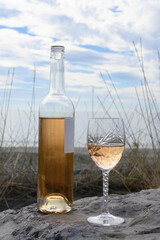 A engraved crystal wine glass, a bottle of rosé wine placed on rocks. The Mediterranean Sea, tall grasses in the background. Camargue, at Saintes-Maries-de-la-Mer. Vertical picture.