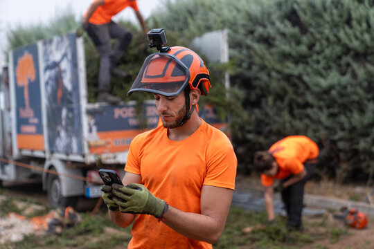 Arborist worker wearing safety helmet with gopro camera checking smartphone during tree pruning operation - Powered by Adobe