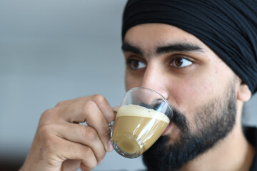 A Young Sikh Man Wearing a Turban Enjoys a Cup of Frothy Coffee or Tea Indoors with a Calm Expression While Looking Away from the Camera
