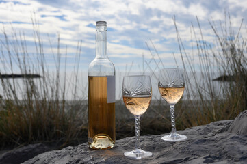 Two engraved crystal wine glasses, a bottle of rosé wine placed on rocks. The Mediterranean Sea, tall grasses and sea walls in the background. Camargue, at Saintes-Maries-de-la-Mer.