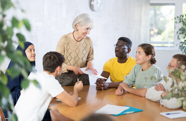 Group of interested people on various ages and nationalities communicating in foreign language speaking club with positive elderly female tutor..