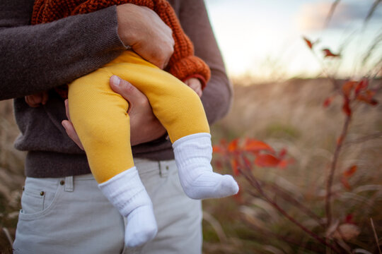 Loving father holding his baby outdoors. Close up of baby's legs. Child wearing fall yellow trousers and brown sweater.