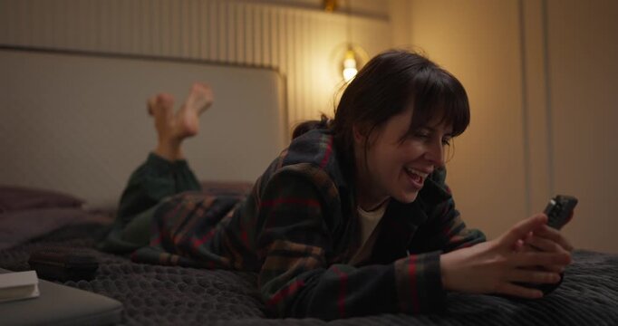 A young woman lies on her bed, relaxed and smiling as she scrolls through her phone in a warm, inviting bedroom