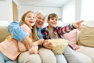 Portrait of an elderly grandmother and grandchildren having fun watching tv together eating popcorn at home