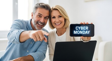 A happy middle-aged couple points at the camera while holding a smartphone displaying "Cyber Monday" and sitting in front of a laptop.