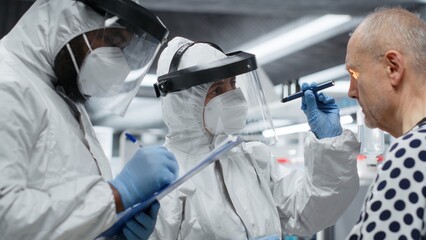 Doctor in isolation examines patient under biohazard conditions, recording treatment outcomes and...