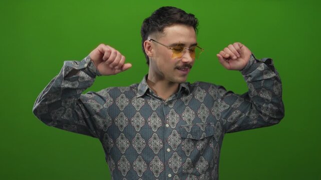 Young hispanic man dancing joyfully against an isolated green background, wearing patterned shirt and unique glasses for a vibrant modern look