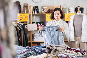 While shopping, Latin American female client looks at products in store and selects jeans trousers. Concept of seasonal wardrobe shopping. Client at shopping time leisure activity.