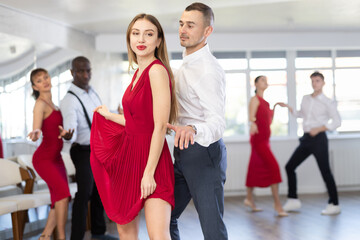 Fototapeta premium Radiant attractive long-haired woman in flowing red dress standing out while learning to dance paired with male partner during dance class. Social dancing concept..