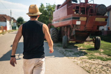 Man wearing straw hat and tank top walking towards agricultural machinery on a sunny day in a village