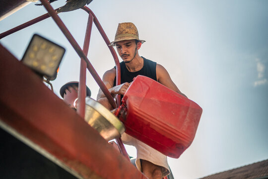 Farmer in straw hat refueling a red agricultural tractor from a jerrycan, ensuring fuel for farming operations