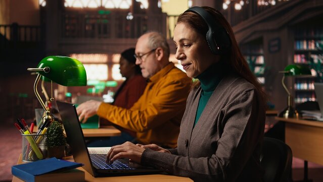 Mature student listening to school webinar on laptop at a library, takes notes and does research during online lecture. Studying remotely with an expert to obtain her diploma. - Powered by Adobe