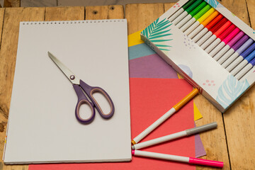 Art and craft supplies arranged on wooden surface, featuring sketchpad, scissors, colored paper,...