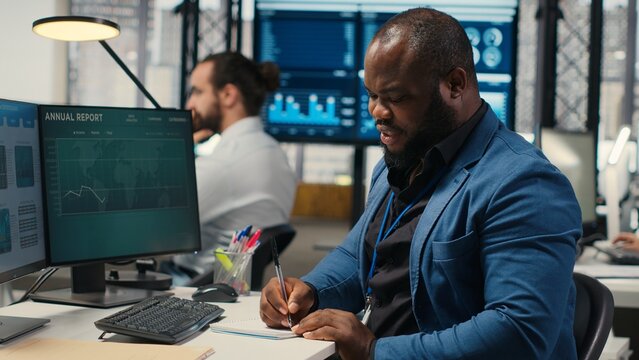Black employee building new development strategy with notes, analyzing data and solutions to forecast project outcomes. Male professional working on briefing reports in the office. Camera B.