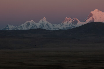 Dramatic sunset view of the Cordillera Huayhuash in Peru. The snow-capped peak of Jerupaja is prominently illuminated by golden alpenglow, rising above the shadowed mountain range.