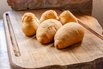 Five pieces of rustic bread with golden crust arranged on wooden surface against stone wall, showcasing traditional homemade baking in cozy kitchen setting.