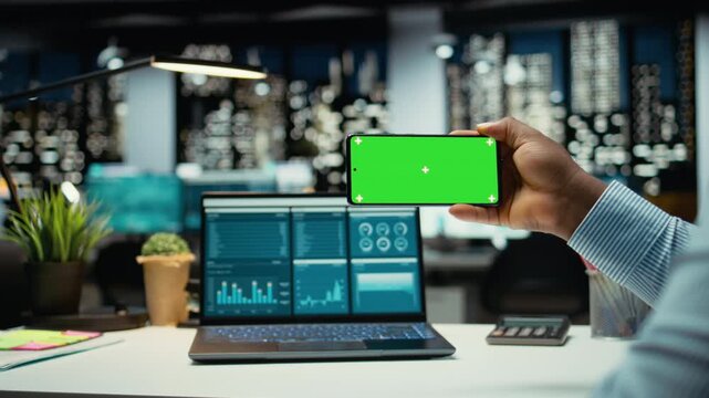 Black office employee working with isolated screen on mobile phone, proofreading and fact checking in the office late at night. Person collecting information on a laptop for monthly report. Camera A.