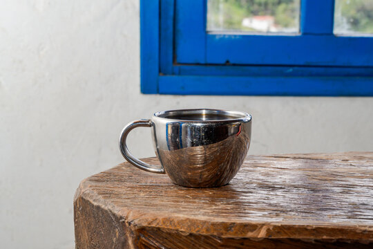 Reflective metal cup on rustic wooden surface near blue-framed window with greenery, capturing cozy and minimalist morning scene with natural light and contrast