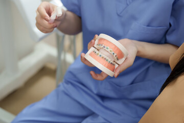 Dental assistant in blue uniform holding orthodontic model with braces and toothbrush explaining...