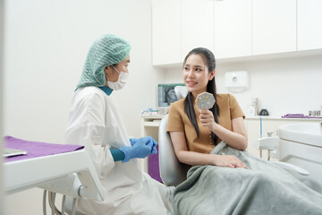 Fototapeta premium Asian dental assistant wearing surgical outfit sitting near female patient in dental chair, discussing procedure while woman holding mirror during preparation for oral treatment in clean clinic