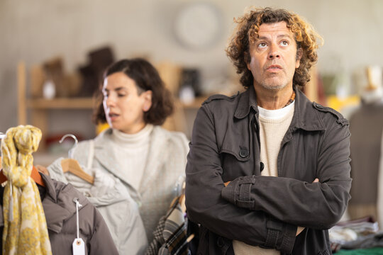 European female customer choose new jacket or coat for winter season, man bored or imitated while waiting for woman in modern clothing store - Powered by Adobe