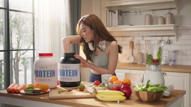 Young Asian woman in sportswear making healthy protein shake smoothie in home kitchen, using organic fresh fruits, vegetables, and whey powder. Wellness, fitness lifestyle, balanced diet, weight loss.