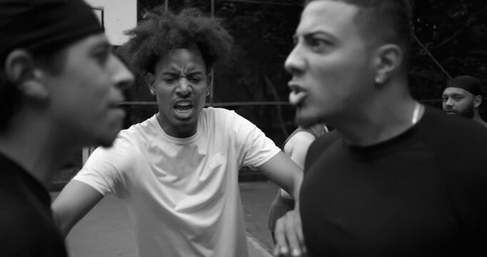 Two men of Latin and African descent arguing face to face on outdoor basketball court, intense confrontation as African descent friend intervenes to calm them, monochrome scene