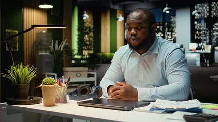 Black male employee closing his lamp and laptop at the office after hours, working late at night to meet deadlines and achieve objectives accomplishment. Doing overtime and strategizing. Camera A.