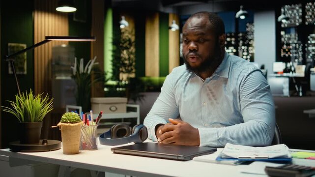 Black male employee closing his lamp and laptop at the office after hours, working late at night to meet deadlines and achieve objectives accomplishment. Doing overtime and strategizing. Camera A.