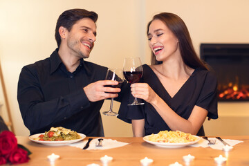 A couple enjoys a romantic dinner, toasting with red wine glasses. They share a table set with delicious dishes, surrounded by soft candlelight and a warm fireplace.