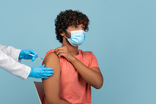 Doctor in gloves gives an intramuscular injection to a male patient wearing a face mask. The patient looks calm while sitting in a chair, during the ongoing COVID-19 pandemic.