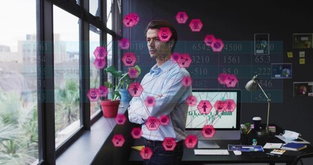 Standing executive wearing blue shirt observing pink data icons near office windows, copy space