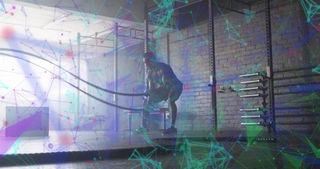 Fototapeta premium Swinging athlete performing battle rope exercise in gym, with metal rig, barbells and weight plates