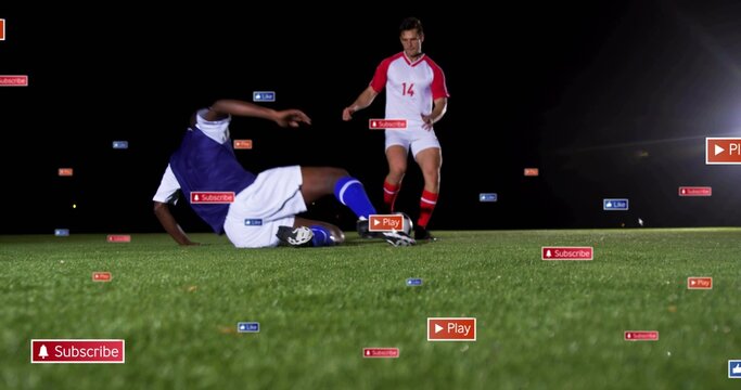 Sliding soccer player in blue kit tackling red opponent on turf under lights, with floating icons - Powered by Adobe