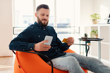A man sits comfortably in a bright room, using his smartphone while holding a credit card. Sunlight streams through large windows, creating a relaxed atmosphere.