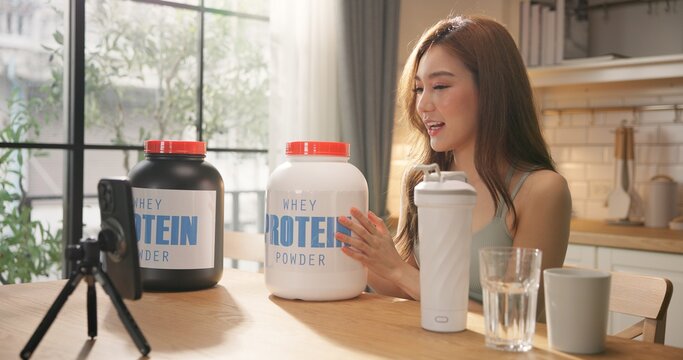 Young Asian female health influencer live streaming on social media sharing nutrition tips with whey protein powder by smartphone. Healthy lifestyle, fitness coaching, clean eating. online marketing.