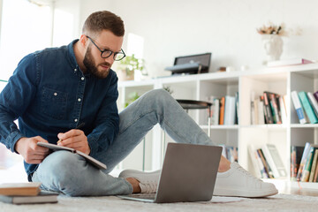 A young man is sitting on the floor in a warm living room, focused on his laptop while taking notes in a notebook. Books are neatly arranged in the background.
