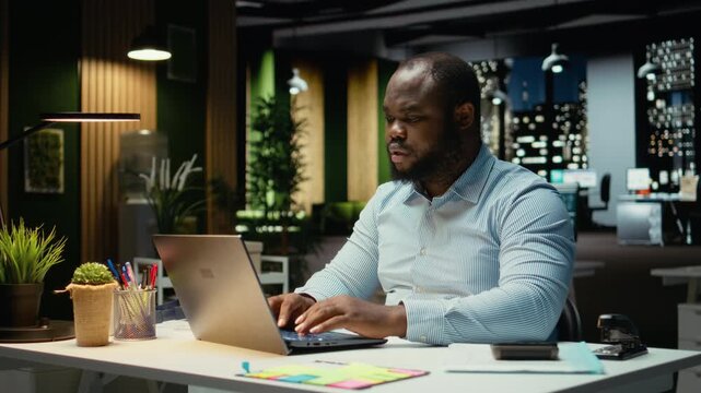 African american guy answering emails and documenting data from online insights for a multinational company, working after hours in a dark office. Enterprise objectives and development. Camera A.