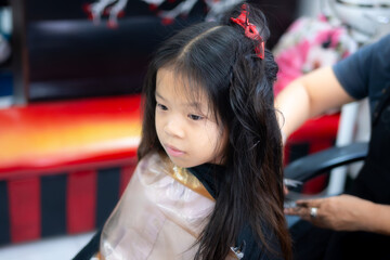 Fototapeta premium A beautiful Asian child girl with long dark hair sits quietly in a salon chair for a haircut. Kid solemn expression and the delicate process of a first haircut or a routine hair maintenance session.