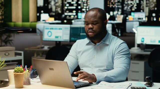 African american employee working at night proofreading web information, writing important data on laptop. Creating a summary and designing strategy to align with enterprise targets. Camera B.
