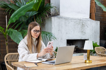 Young woman working remotely with smartphone and laptop at an outdoor cafe
