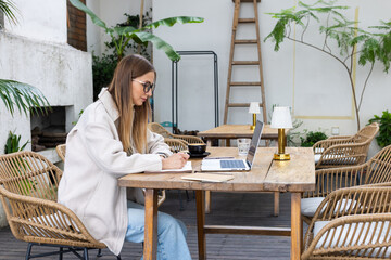 Young woman working on laptop and writing in notebook at outdoor cafe