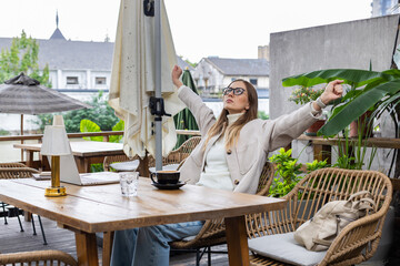 Young woman stretching at an outdoor cafe while working on a laptop