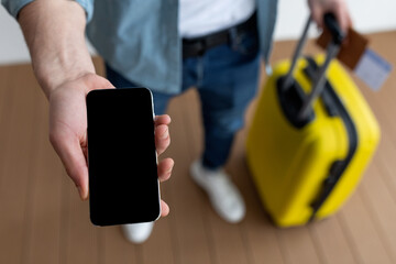 Person stands in a bright indoor area, holding a smartphone in one hand and a yellow suitcase...