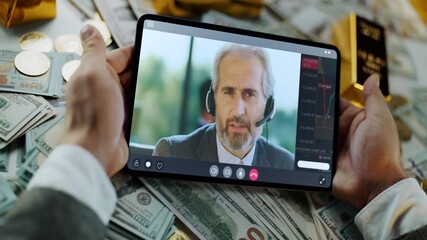 Successful businessman holding a digital tablet and having an online video conference with a stock market analyst over a table covered with dollar bills, gold coins, and bullion bars - Powered by Adobe