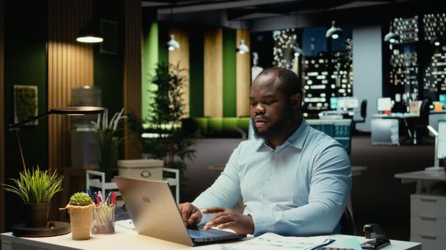 African american employee working at night proofreading web information, writing important data on laptop. Creating a summary and designing strategy to align with enterprise targets. Camera A.