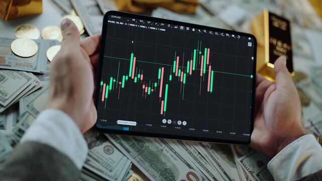 Successful businessman holding a tablet showing a candlestick chart for stock market trading, surrounded by a pile of cash, gold coins, and bullion bars representing wealth and investment