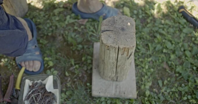 Close-up shot showing a person's hands engaged in manual work outdoors. They are meticulously bending and straightening a piece of wire using pliers and a hammer against a wooden block.