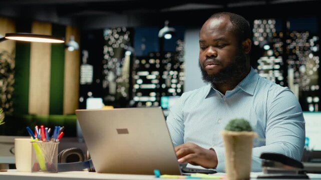 Black employee reading emails and documenting data from web insights for a multinational company, working after hours in a dark office. Enterprise objectives and development. Camera B.