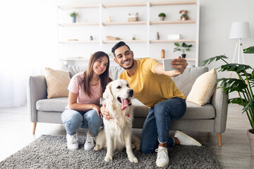 A young couple sits on a soft rug in a comfortable living room, smiling as they take a selfie with their friendly golden retriever. The atmosphere feels warm and inviting.